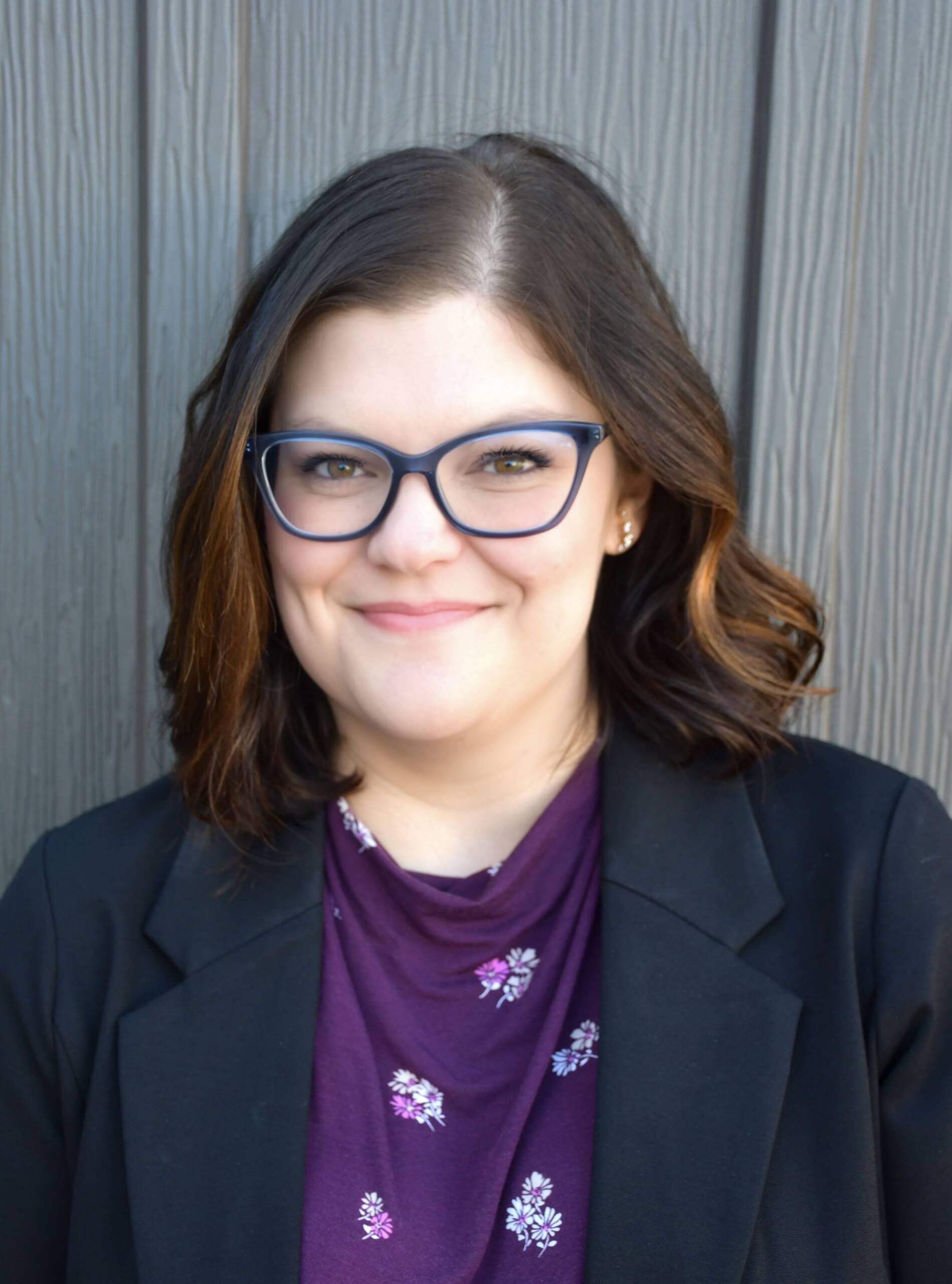 A person with shoulder-length brown hair, wearing blue-rimmed glasses, a dark blazer, and a purple top with floral patterns is seen standing in front of a vertically paneled wooden wall, smiling at the camera.
