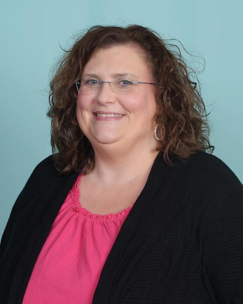 A woman with shoulder-length curly hair, wearing glasses, a black cardigan, and a pink top, smiles against a plain teal background.