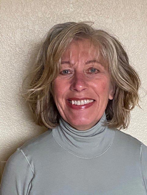 A middle-aged person with short, wavy hair smiles warmly at the camera. They are wearing a light gray turtleneck sweater. The background is a plain off-white wall.