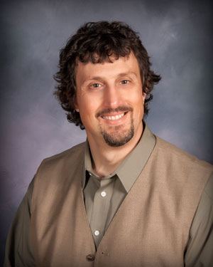 A man with curly dark hair and a goatee is smiling at the camera. He is wearing a sage green shirt with a light brown vest. The background is a softly blurred gradient of blue and purple tones.
