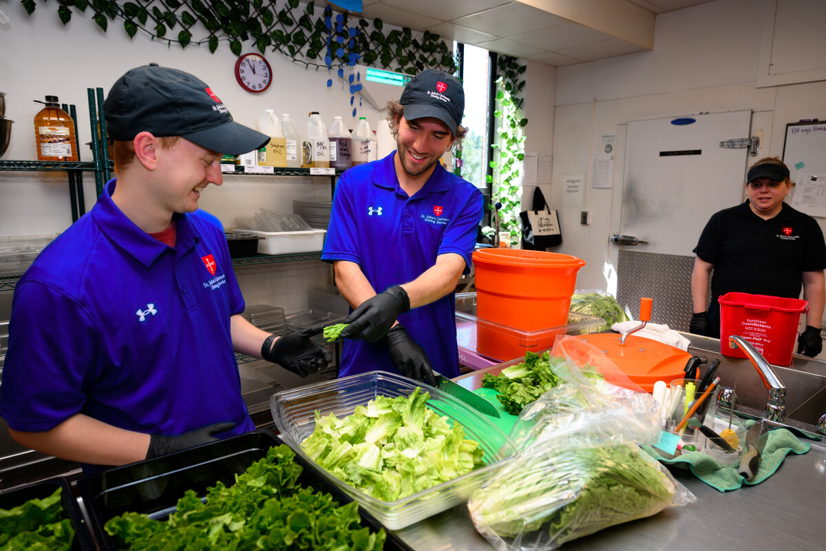 Three people are preparing fresh green lettuce in a well-organized kitchen. Two individuals in matching purple shirts, black gloves, and hats are working together, while a third person in a black shirt stands by and observes. The kitchen appears clean with ample supplies.