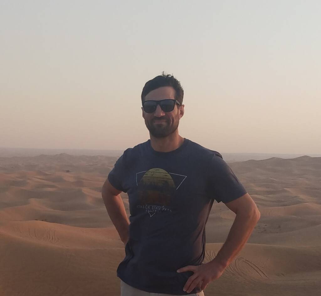 Current image: A person with short dark hair, wearing sunglasses and a dark t-shirt, stands with hands on hips in a desert landscape with rolling sand dunes under a clear sky. The sun is either rising or setting, casting a warm glow over the scene.
