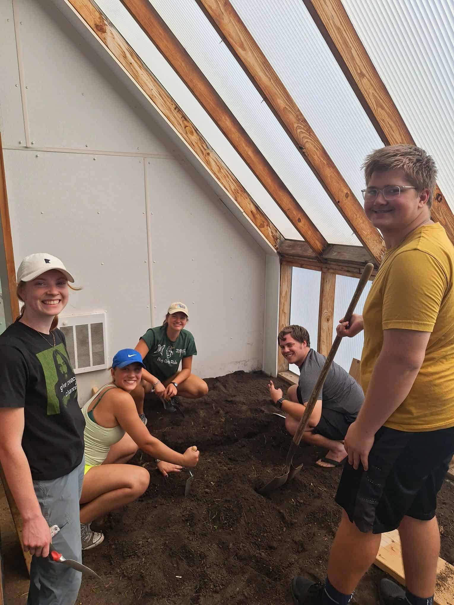 A group of five people smiling and working together in a garden greenhouse. Four of them are kneeling and using small tools to work the soil, while one stands holding a large garden fork. The greenhouse has wooden beams and translucent walls.