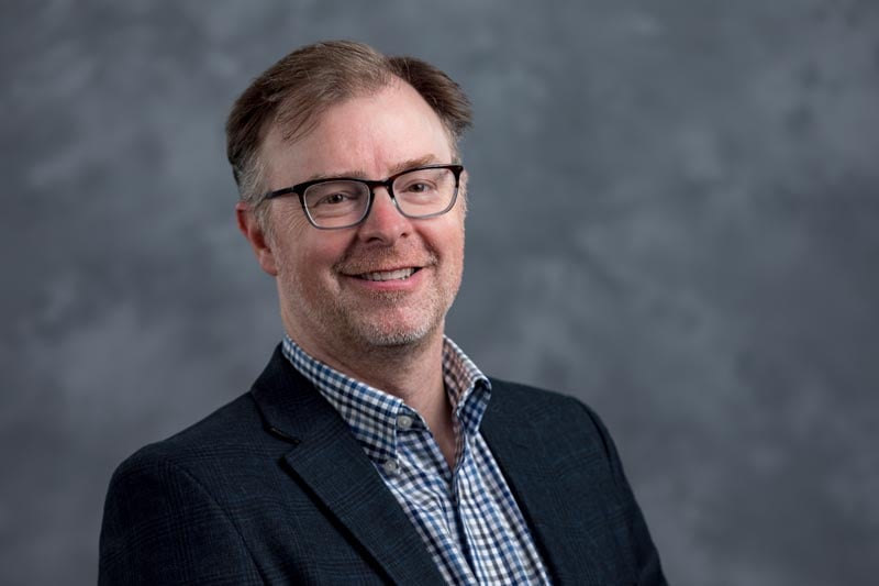 A man with short, light brown hair and glasses smiles at the camera. He is wearing a dark blazer over a checkered shirt. The background is a simple gray, giving the image a professional and polished look.
