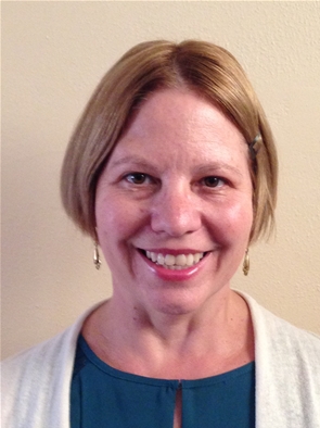 A smiling person with short light brown hair, accessorized with a small clip on the left side. They are wearing a blue top under a light-colored cardigan and small dangling earrings. The background is a plain beige wall.