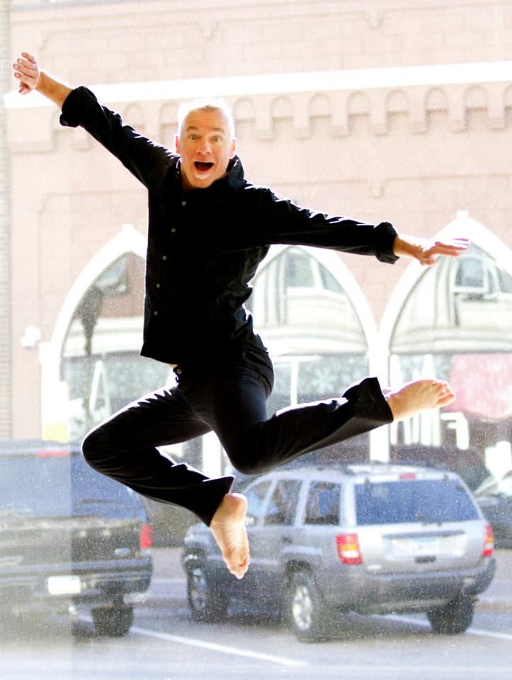 Person with short light hair, dressed in all black, mid-air in a joyful leap with arms and legs extended. Background includes large windows with an urban street scene, featuring cars and a building with arched windows.