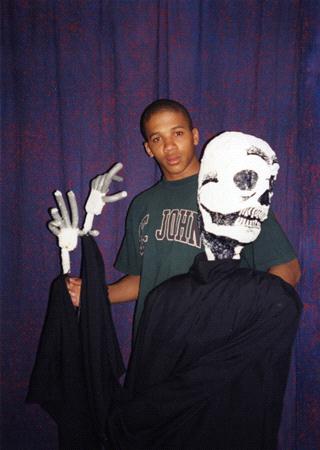 A person wearing a St. John's T-shirt poses with a large skeleton puppet dressed in black. The skeleton has white gloves and a grinning skull. They stand in front of a dark blue curtain.