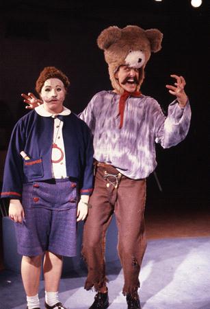 A person dressed as a clown with white face paint and a blue outfit stands next to another person wearing a large teddy bear headpiece and long-sleeve shirt, appearing animated with a playful gesture. The background is dimly lit.