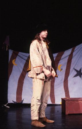 A person in a fringed outfit and black hat stands on stage, set against a backdrop with red and white stripes and star and moon designs. A small chest is on the floor beside them.