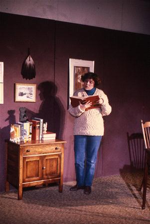 A person in a heavy sweater stands reading a book beside a wooden cabinet with books and small decorations. The room has a dark purple wall, framed pictures, and a wooden chair, creating a cozy setting.