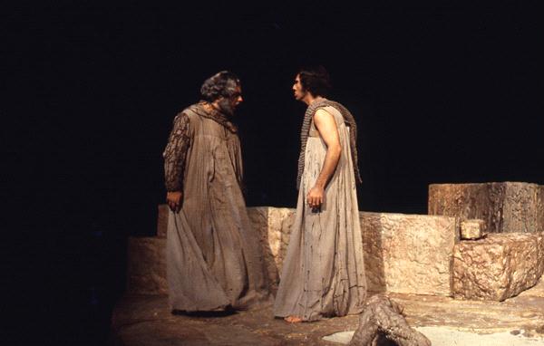 Two actors on stage dressed in ancient-style robes engage in a dramatic scene. They stand on a rocky, uneven surface with dim lighting, evoking a classical or mythological atmosphere.