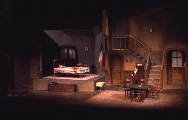 A dimly lit stage set depicts a rustic room with an unmade bed, wooden table, and chairs. A staircase leads to a balcony above. The setting has a worn, vintage feel, with an Irish flag hanging on the wall.