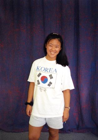 Person smiling and posing in front of a dark curtain backdrop, wearing a white T-shirt with the word "Korea" and the South Korean flag printed on it. They have dark hair and are wearing light-colored shorts.
