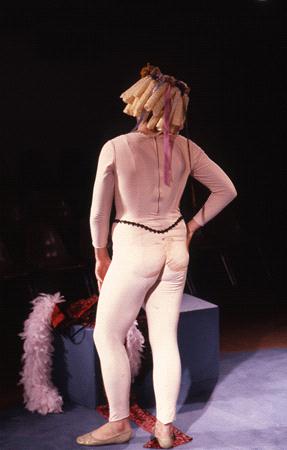 A person dressed in a cream-colored bodysuit with a wig made of rolled papers stands on stage with their back facing the camera. A pink feather boa and patterned fabric are on the floor nearby. The background is dimly lit.