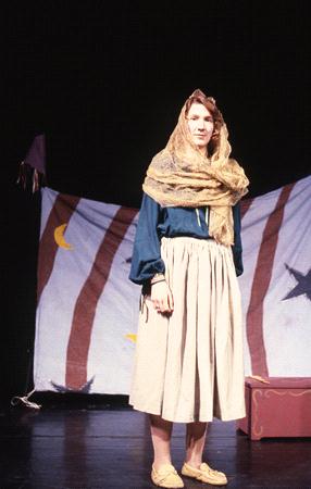 A person stands on stage wearing a headscarf and long skirt, with a backdrop featuring stars and moons on striped fabric.