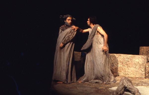 Two people are dressed in ancient-style robes on a dimly lit stage, with one reaching out to the other. The setting appears to be a dramatic scene with stone-like structures in the background.