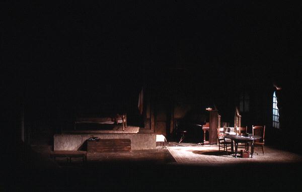 Dimly lit stage set featuring a dark room with a bed, table, chairs, and a small couch. Soft light filters through a window, highlighting portions of the furniture and casting shadows.