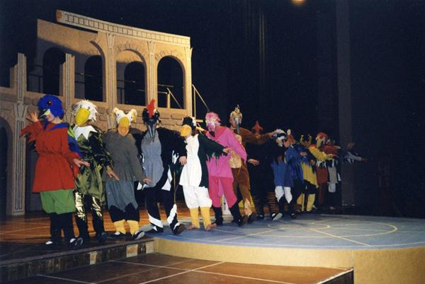 A group of people on stage dressed in colorful bird costumes, performing in front of a set resembling ancient architecture. The scene is well-lit, showcasing a variety of bright outfits and headpieces.