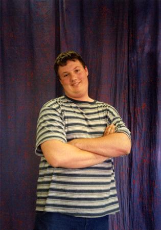 A person wearing a striped shirt stands with arms crossed, smiling in front of a dark blue curtain backdrop.