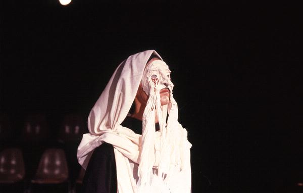 A person wearing a white cloth draped over their head and face stands against a dark background. The fabric is layered, with some parts falling over their eyes and extending downwards.