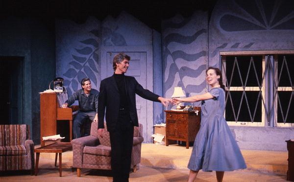 A man and a woman dance on stage in a set designed as a living room. Another man sits at a piano, smiling at them. The room has painted walls with abstract patterns, a window, and furniture including chairs and a lamp.