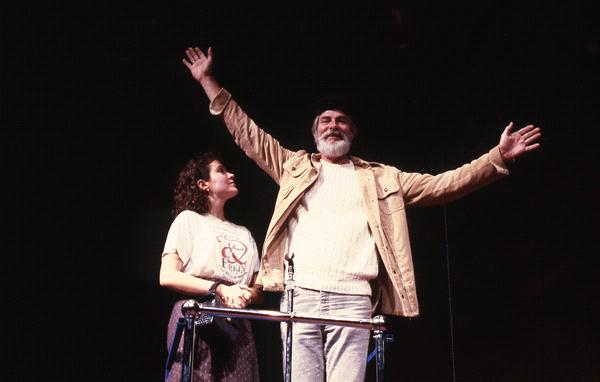 A man with a white beard and a beanie raises his arms triumphantly. A woman with curly hair stands beside him, looking up, holding onto a railing. She is wearing a T-shirt with a graphic. The background is dark.