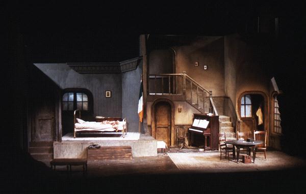 A dimly lit theater stage set depicting an interior room. Features include a raised bed, an upright piano with sheet music, a table with chairs, a staircase leading to an upper level, and muted walls with minimal decor.