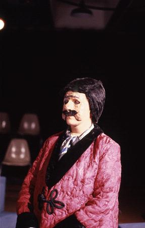 A person in a pink coat with black trim and a white cravat stands in a dark room. They are wearing theatrical makeup with an exaggerated mustache and a mask-like appearance. Empty beige chairs are visible in the background.