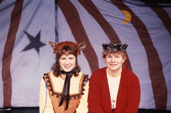 Two people smiling in front of a backdrop with a star and crescent moon. One wears a costume with animal ears and a brown outfit, while the other wears a red sweater and a decorative crown.