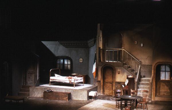 Stage set depicting a dimly lit, rustic interior with a bed, a table and chairs, and a staircase. An Irish flag is draped over a rail, suggesting an old Irish home. The scene conveys a vintage or historical atmosphere.