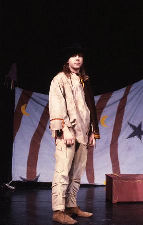A person wearing a brown coat, a scarf, and a black hat stands on a stage. Behind them is a backdrop with red and white stripes, and moon and star symbols. A red box is on the stage floor.