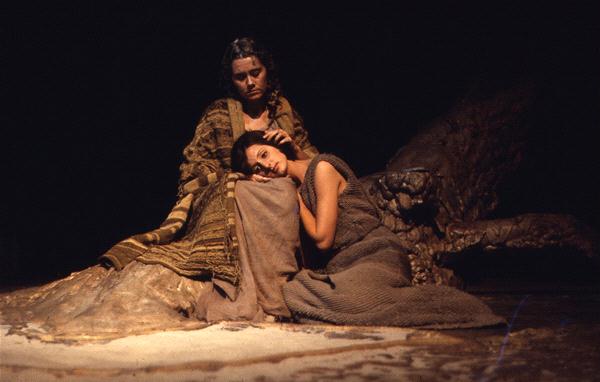 Two women sit in dim lighting on a textured, earthy surface. One woman sits upright, wearing a patterned shawl, while the other rests her head on her lap, wearing a plain, sleeveless dress. The mood is somber and contemplative.