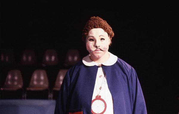 A person dressed in a blue outfit with a white collar and a large necklace shaped like a clock stands against a dark background. They are wearing a theatrical mask with exaggerated features and a reddish-brown wig.
