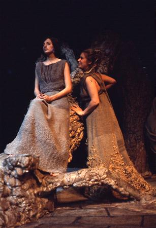 Two women in medieval-style costumes stand on a rocky, stage-like setting. One woman sits with hands clasped, while the other stands leaning on a tree, both gazing towards the same direction, illuminated by soft stage lighting.