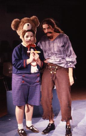 Two people in theatrical costumes stand together. One wears a blue and red outfit with a large teddy bear headpiece and holds a banana. The other wears a distressed pirate-like outfit with an exaggerated mustache.