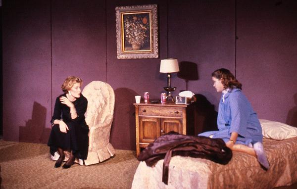 Two women are in a room with purple walls and a floral painting. One woman is seated in a patterned armchair, while the other sits on a bed. A bedside table with a lamp and items is between them. The setting suggests a conversation.