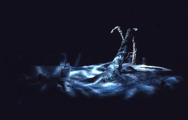 Dimly lit stage scene depicting a surreal, barren landscape with a twisted tree or structure covered in snow. The setting is dark and moody, evoking a sense of solitude and stillness. Subtle shadows suggest an ethereal, mystical atmosphere.