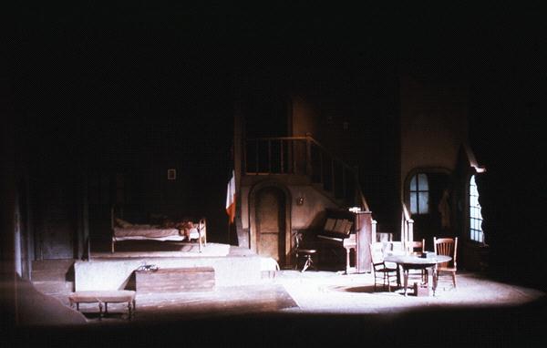 Dimly lit stage set depicting a rustic room with a wooden staircase, a piano, and a round table with chairs. There is a bed with a draped blanket, and a soft, warm glow suggests an old-world, cozy atmosphere.