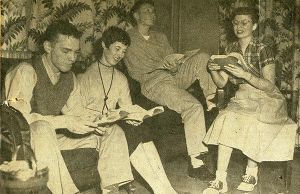 Four people sitting and reading books in a room with patterned wallpaper. Three are on a couch, one sits on the armrest. They appear relaxed and engaged, with casual 1950s attire.