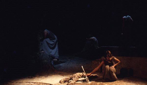 A dimly lit stage scene with two figures in gray garments. One figure is kneeling beside a large root or branch, while another sits on a platform in the background. The atmosphere is somber and theatrical.