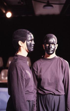Two individuals wearing black clothing and masks painted with metallic black paint stand in dim lighting. The background is blurry and a row of empty chairs is visible.