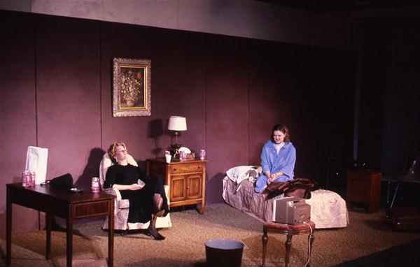 Two people are in a dimly lit room. One sits on a chair dressed in black, while the other is seated on a bed wearing blue. The room includes a wooden table, lamp, nightstand, TV, and wall art.