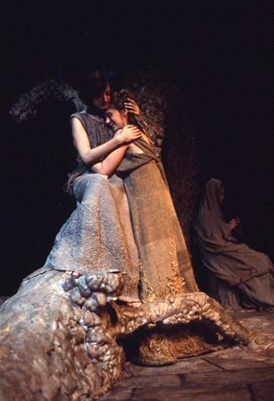 Two people in gray, textured costumes sit closely together in a dimly lit, rocky setting. One person comforts the other, with an embrace and their head resting on the other's shoulder. A third figure sits in the background, partially covered.