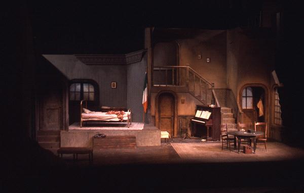 Dimly lit stage set of a modest room with a bed, piano, table, and chairs. Stairs lead to an upper level. An Irish flag hangs near the piano. The scene has an atmospheric, rustic feel with muted colors and shadows.