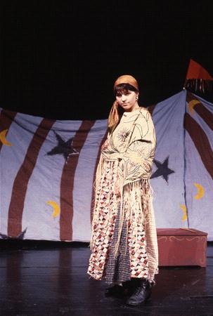A person stands on a stage wearing a patterned long dress and headscarf. The backdrop features stripes with crescent moons and stars. A red box is on the stage, and the lighting suggests a theatrical performance.