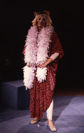 Person in a theatrical costume with a red patterned robe, white leggings, and ballet slippers, adorned with a pink feather boa and an animal-like mask, standing on a blue stage under dramatic lighting.