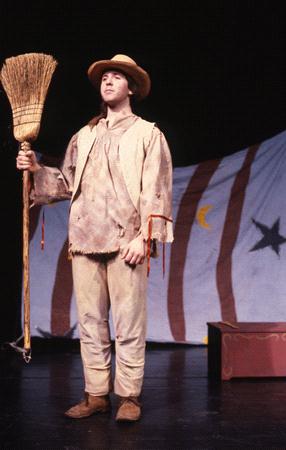 A person in a rustic costume holds a broom on stage. They wear a wide-brimmed hat and simple, loose clothing. Behind them, a backdrop features vertical stripes and star and moon symbols. A wooden box sits nearby.
