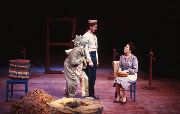 A theatrical scene features a person in a dragon costume crouching beside a standing person in a uniform, both facing a seated person holding a basket. The stage includes two chairs, a rope border, and a backdrop door frame.