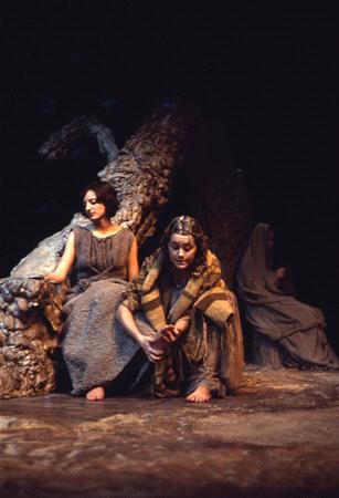 Three people in ancient-style costumes sit on a rocky stage. The two in the foreground are in dramatic poses, with one looking away while the other gazes downwards. A third person sits in the background, partially obscured by shadows.