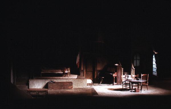 Dimly lit stage set depicting a rustic room with wooden furniture. A round table with chairs is at the center, a bed is to the left, and a piano to the right. Draped curtains and low lighting add an intimate, dramatic atmosphere.
